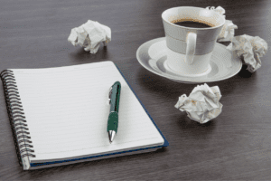 Um caderno em branco ao lado de uma xícara de café e alguns papeis amassados.